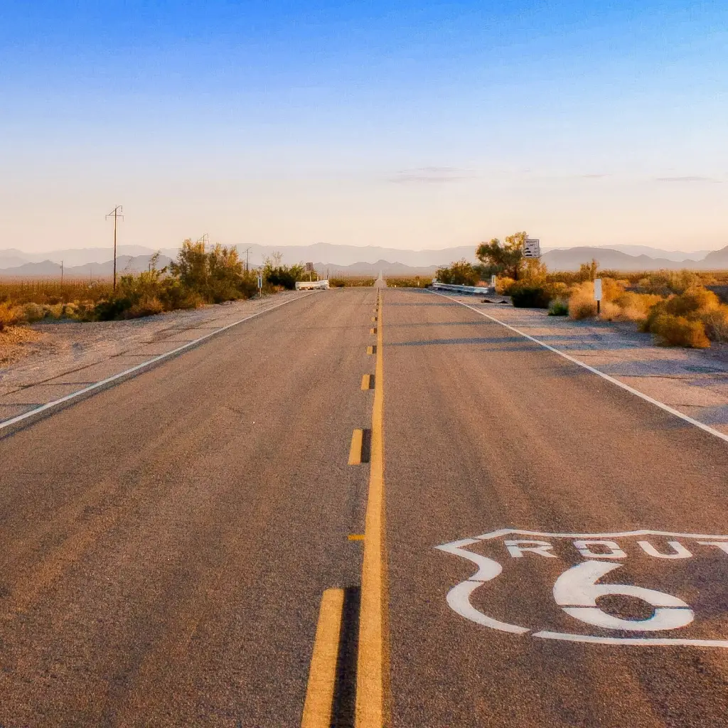 Eine leere Strecke der Route 66, die bei Sonnenaufgang durch die Wüste führt, mit langen Schatten und fernen Bergsilhouetten.