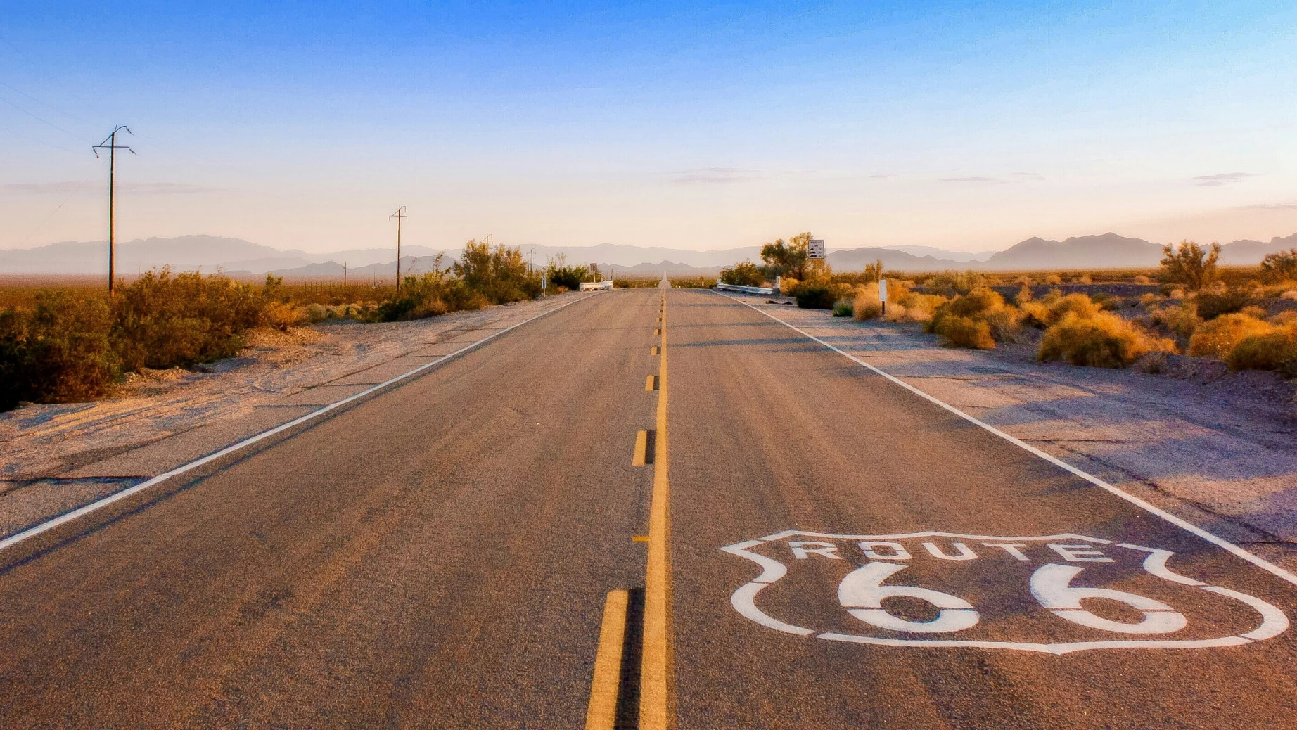 Eine leere Strecke der Route 66, die bei Sonnenaufgang durch die Wüste führt, mit langen Schatten und fernen Bergsilhouetten.