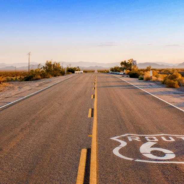 Eine leere Strecke der Route 66, die bei Sonnenaufgang durch die Wüste führt, mit langen Schatten und fernen Bergsilhouetten.