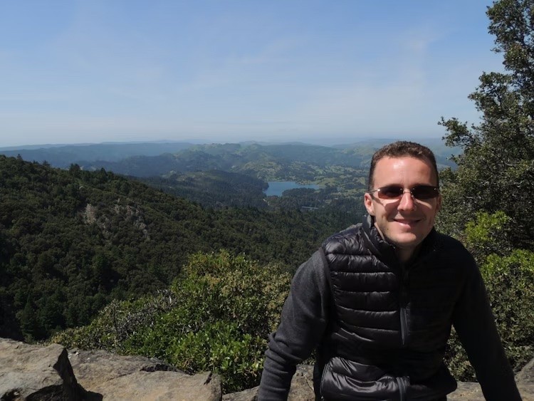 Mann mit Sonnenbrille sitzt auf Steinen vor Tal mit See