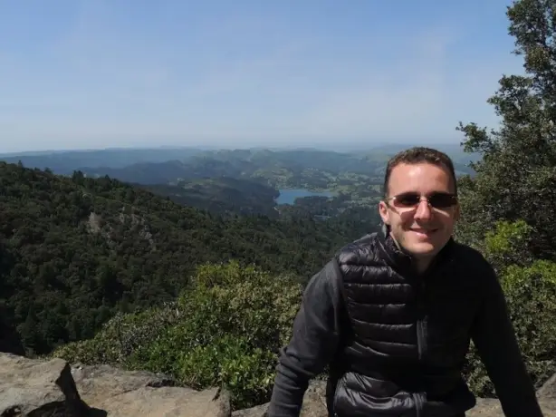 Mann mit Sonnenbrille sitzt auf Steinen vor Tal mit See