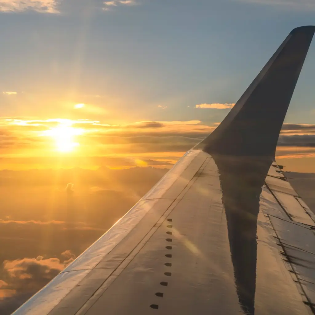 Blick aus dem Fenster eines Flugzeugs bei Sonnenaufgang, mit goldenem Licht über den Wolken und dem Flugzeugflügel, der sich in den Himmel reckt.