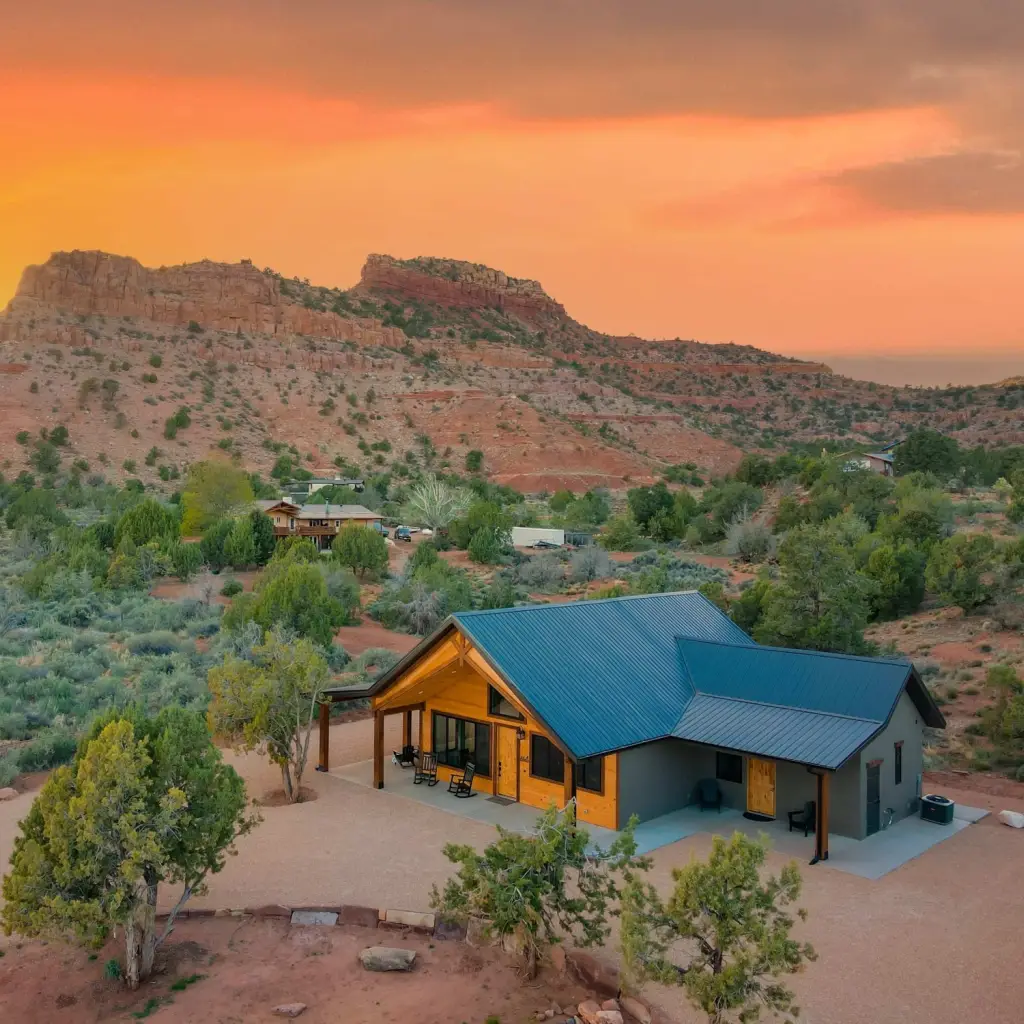 Luftaufnahme der Casa Vermillion, einer modernen Hütte mit türkisfarbenem Metalldach und überdachter Veranda, inmitten von Wüstenbuschland mit dramatischen roten Felsformationen und einem leuchtend orangefarbenen Abendhimmel nahe Kanab, Utah, USA.