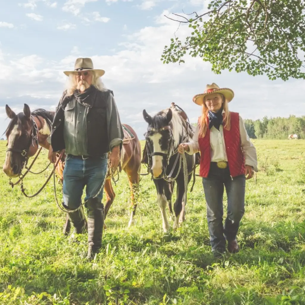 Mann und Frau in Cowboy-Kluft spazieren mit zwei Pferden über Wiese