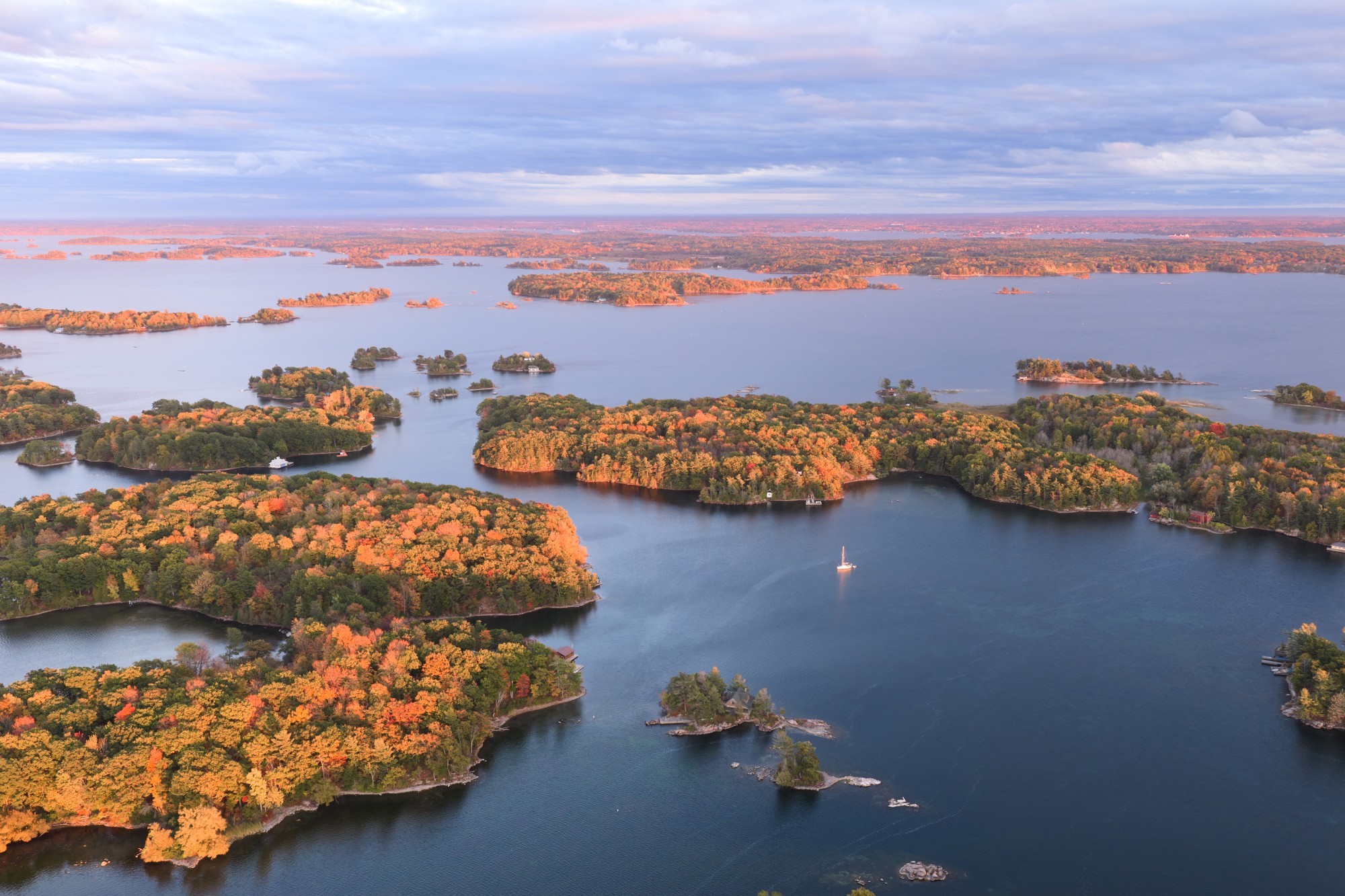 Vogelperspectief over Thousand Islands in Oost-Canada. Tegen zonsondergang aan, met blauwe water en rood-oranje verkleurende bomen. Op het water vaart een zeilboot.