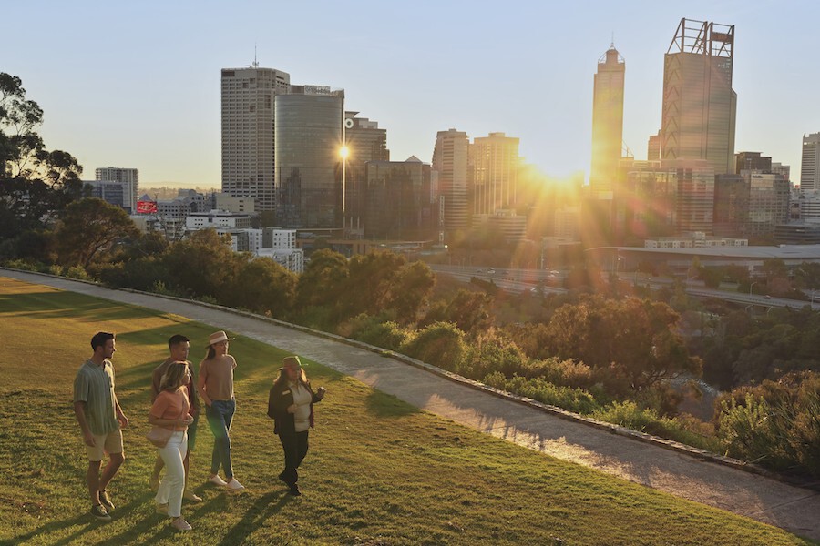 Eine Gruppe von Menschen, die im King's Park in Perth spazieren gehen, während die Sonne zwischen den Gebäuden der Stadt im Hintergrund scheint, in Australien