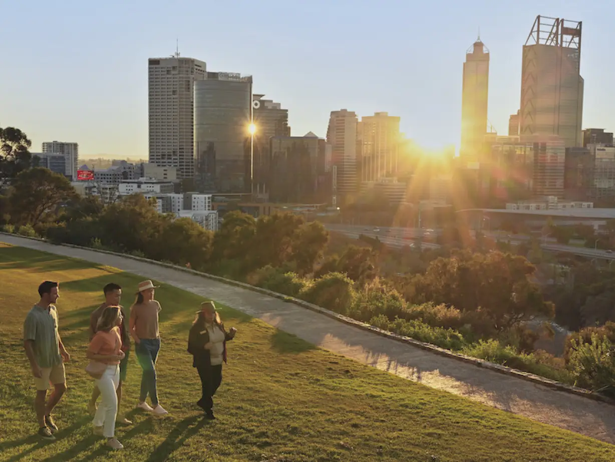 Eine Gruppe von Menschen, die im King's Park in Perth spazieren gehen, während die Sonne zwischen den Gebäuden der Stadt im Hintergrund scheint, in Australien