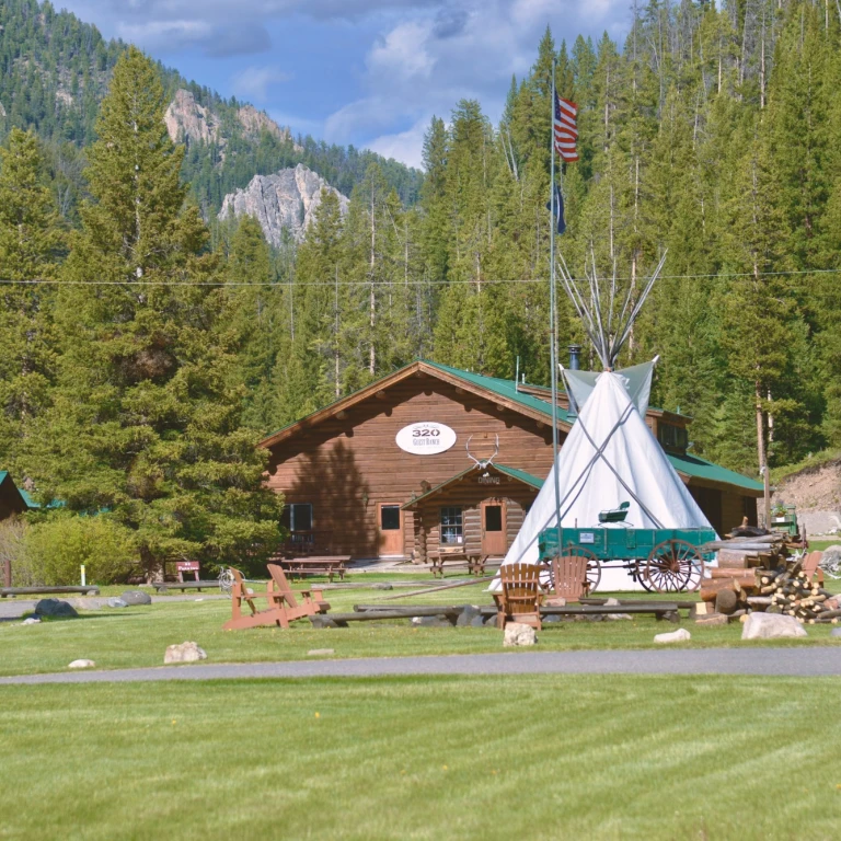 Das Hauptgebäude der 320 Guest Ranch mit einem großen Tipi, einem historischen Planwagen, Adirondack-Stühlen und Blockhütten auf einer grünen Rasenfläche, umgeben von Nadelwald und felsigen Berggipfeln nahe West Yellowstone, Montana, USA.